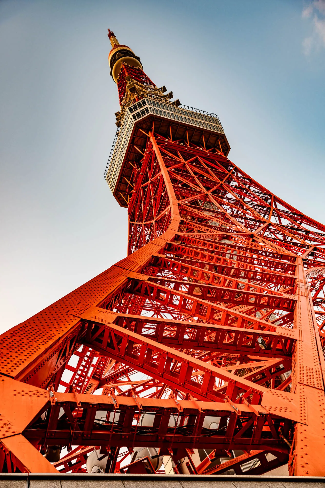 This image is a low angle shot of the Tokyo Tower, a large red, lattice-style tower, with the sky in the background. The tower is made of many interconnected pieces of metal and is a popular landmark in Tokyo, Japan. The tower looks like a triangular prism or a pyramid, as the sides slope in toward each other. The lower portion of the tower is visible with the tower extending far into the sky. The tower is a bright orangey red color. The top of the tower is not visible in the image. The sky is light blue with a faint white cloud in the upper right corner.