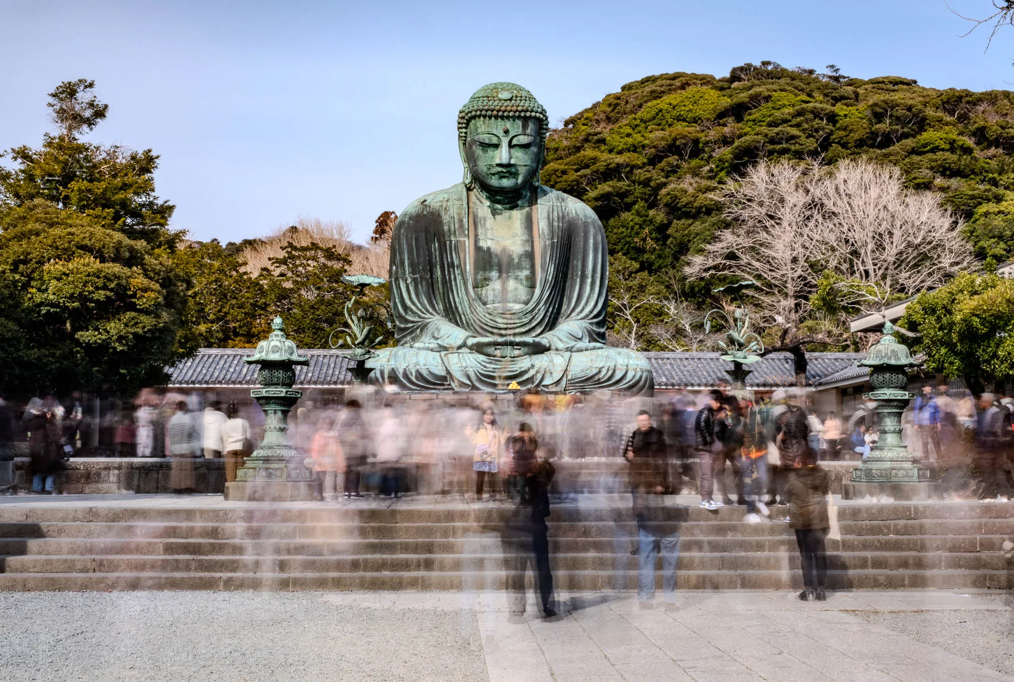 The image shows a large, bronze Buddha statue sitting with his legs crossed, hands in his lap, in front of a temple. The statue is green with age and has a serene expression on its face. The statue is surrounded by people, who are blurred as they walk by. The Buddha statue is seated in a large courtyard. There are some small, ornate structures in the background, as well as trees.  The sky is a pale blue with a few wispy clouds. There are stone steps in the foreground. The image is taken from a low angle, looking up at the Buddha statue.