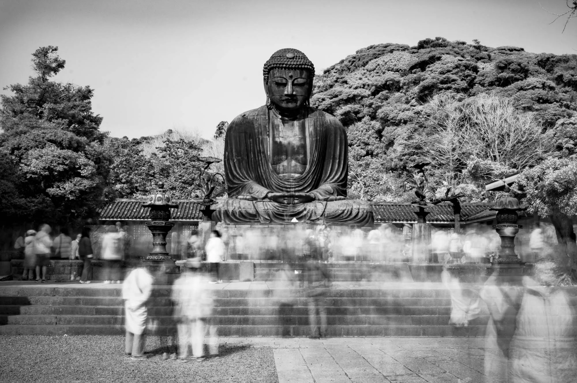 The image is a black and white photograph of a large Buddha statue. The statue is sitting in a meditative pose, with its hands resting in its lap. The statue is made of stone and is very detailed. The Buddha is wearing a robe, and it has a calm and serene expression on its face. The statue is surrounded by trees and a stone structure. There are also many people in the image, but they are blurred and difficult to make out.  The people are in the foreground of the image and they are all moving around the statue. The photo is taken from a low angle, looking up at the statue.  The overall feel of the image is one of peace and tranquility.