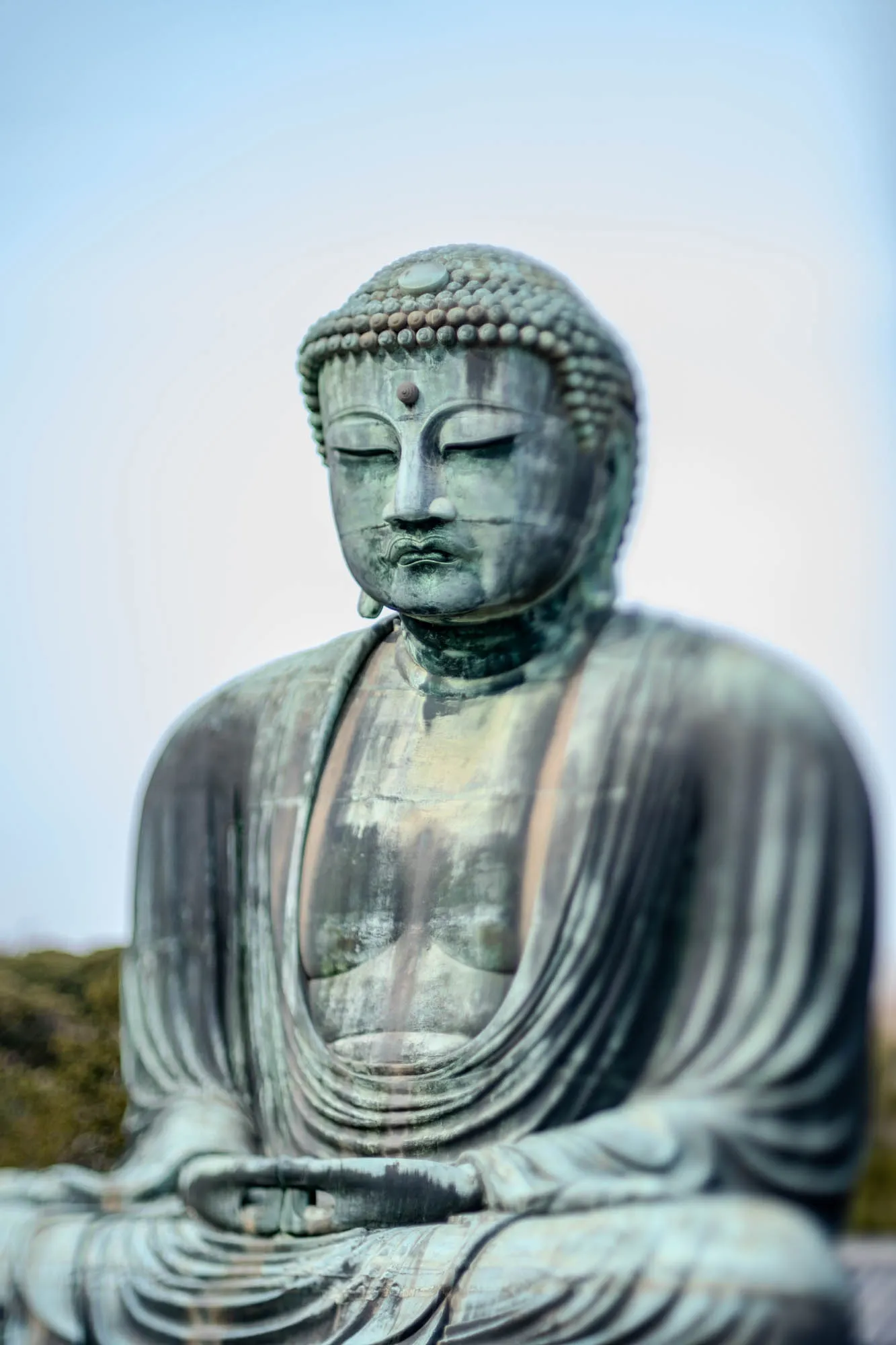 The image is a close-up shot of a large, bronze statue of Buddha sitting in a meditative pose with his eyes closed. The statue is very detailed, and the face of the Buddha is visible, with a calm expression. The Buddha has a small dot on his forehead and a small curl of hair on each ear. He is wearing robes that are folded over his legs. The background is out of focus but appears to be a light blue sky.