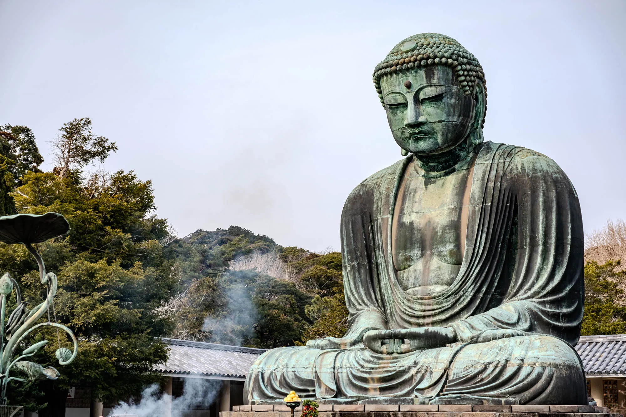 A large statue of Buddha sits in a meditative pose against a clear, light blue sky. The statue is made of bronze and has a greenish patina. The Buddha is wearing a robe that falls to his feet. His hands rest in his lap in the traditional mudra pose, with his fingertips touching. His face is serene and peaceful, with closed eyes. The statue is set on a low platform and is surrounded by green trees.  In front of the statue is a small incense burner. The smoke from the burner can be seen rising up. There are also some yellow fruits on a small platform, which are a symbol of offerings. The Buddha statue is a very impressive sight, and it inspires a sense of peace and tranquility.