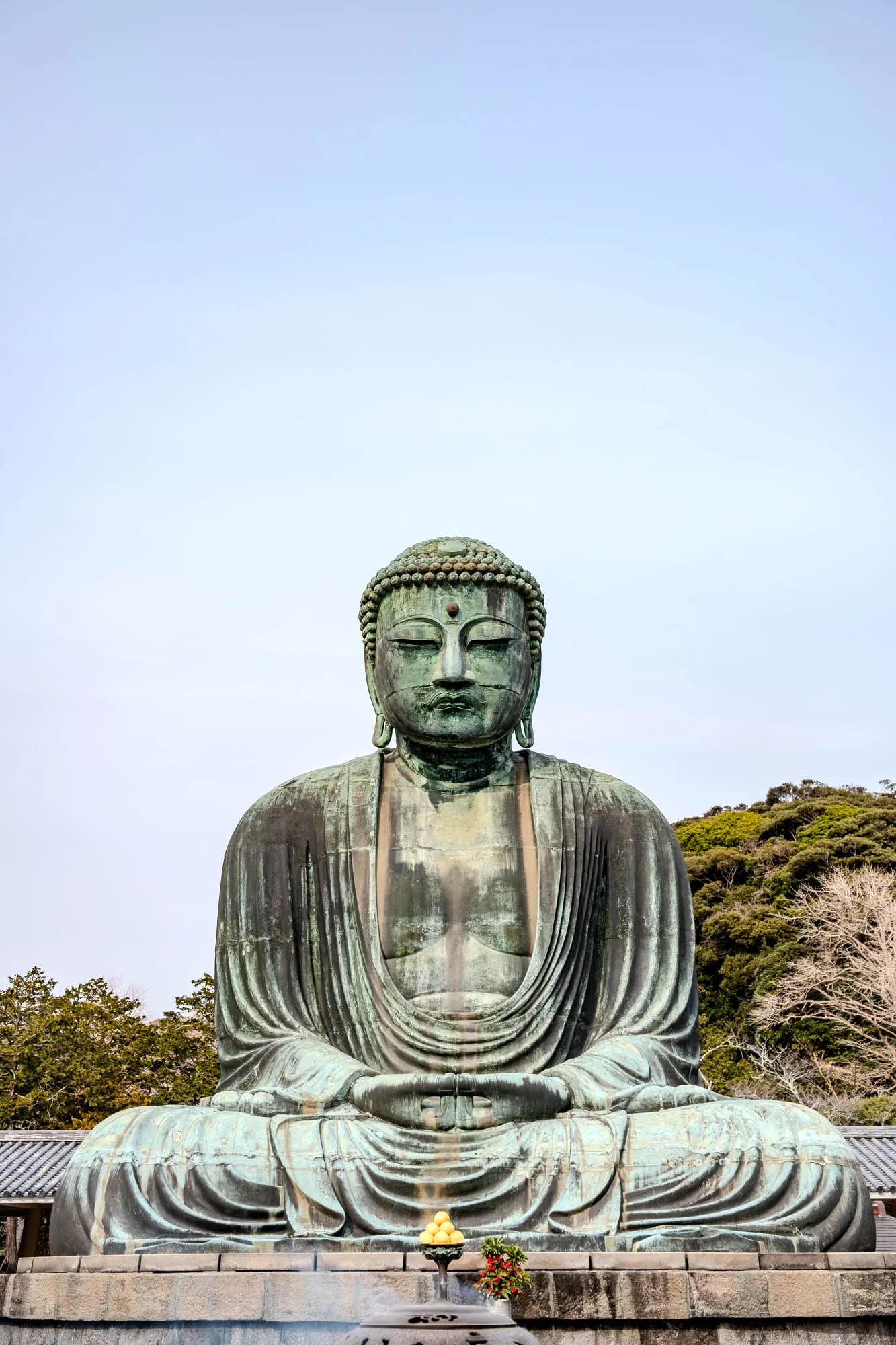 The image depicts a large, bronze statue of the Buddha, seated in a meditative pose with his hands resting in his lap. The statue is weathered and has a greenish patina. The Buddha is wearing a robe that falls to his feet in folds.  The statue is set against a clear blue sky with trees in the background. At the base of the statue, there is a small offering of fruit, a small incense burner, and flowers. The statue appears serene and peaceful.  The image conveys a sense of tranquility and spiritual devotion.