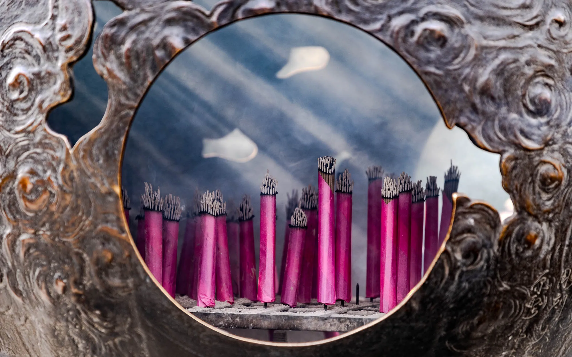 The image shows a close-up of a round, carved, metal object with a circular opening in the center. Inside the opening, there are a number of tall, thin incense sticks standing upright in a row. The sticks are a deep reddish-pink and have a dark gray ash at the top, with a faint plume of smoke rising from them. The background of the opening is out of focus and appears as a hazy, gray-blue with a faint, white, blurry shape in the top center. The front of the metal object is carved with a swirling, textured pattern and appears to be made of a dark, metallic material. The texture of the object is rough and uneven, with a slightly grainy appearance.