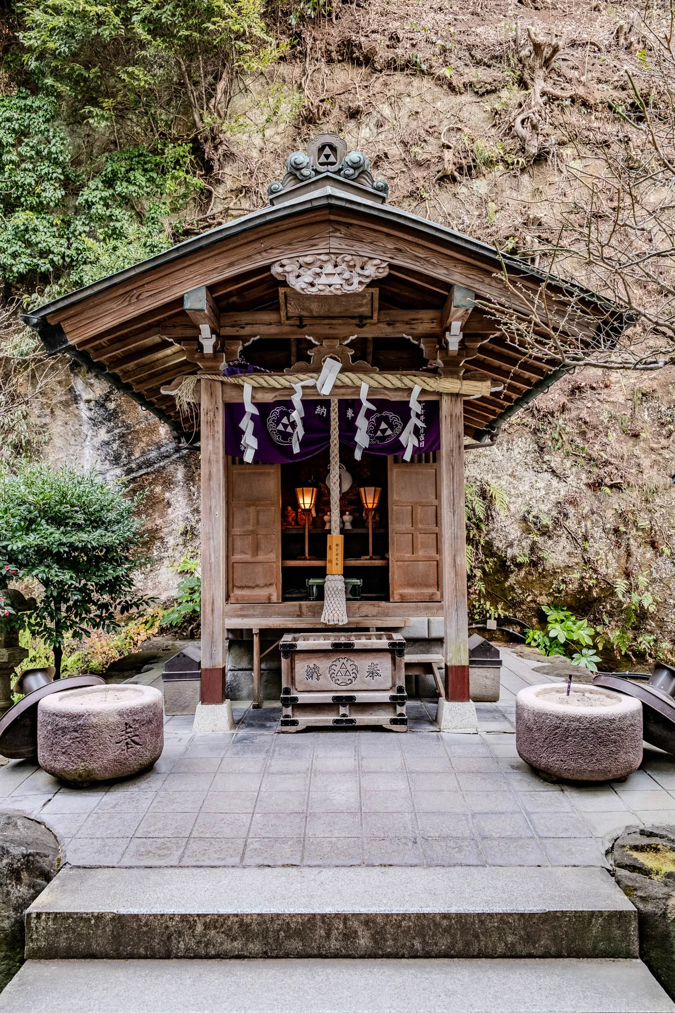 This is a photo of a small, wooden shrine set into a rocky cliff.  The shrine is made of weathered, dark wood and has a gently sloping roof with a decorative, triangular motif on the front. It is decorated with a dark purple fabric draped over the opening, tied with a thick rope and many pieces of white paper tied with colorful ribbons, which hang down.  Two lanterns are lit inside the shrine, and you can see a decorative, white tassel dangling from a rope inside. The shrine is elevated on a stone platform, and the platform has a large, wooden chest with a decorative motif on it, a few stone steps lead down from the front of the shrine and  there are two stone basins in front of it, one on either side. The cliff face behind the shrine is covered in green plants and moss, and the background is filled with green leaves and the branches of trees.