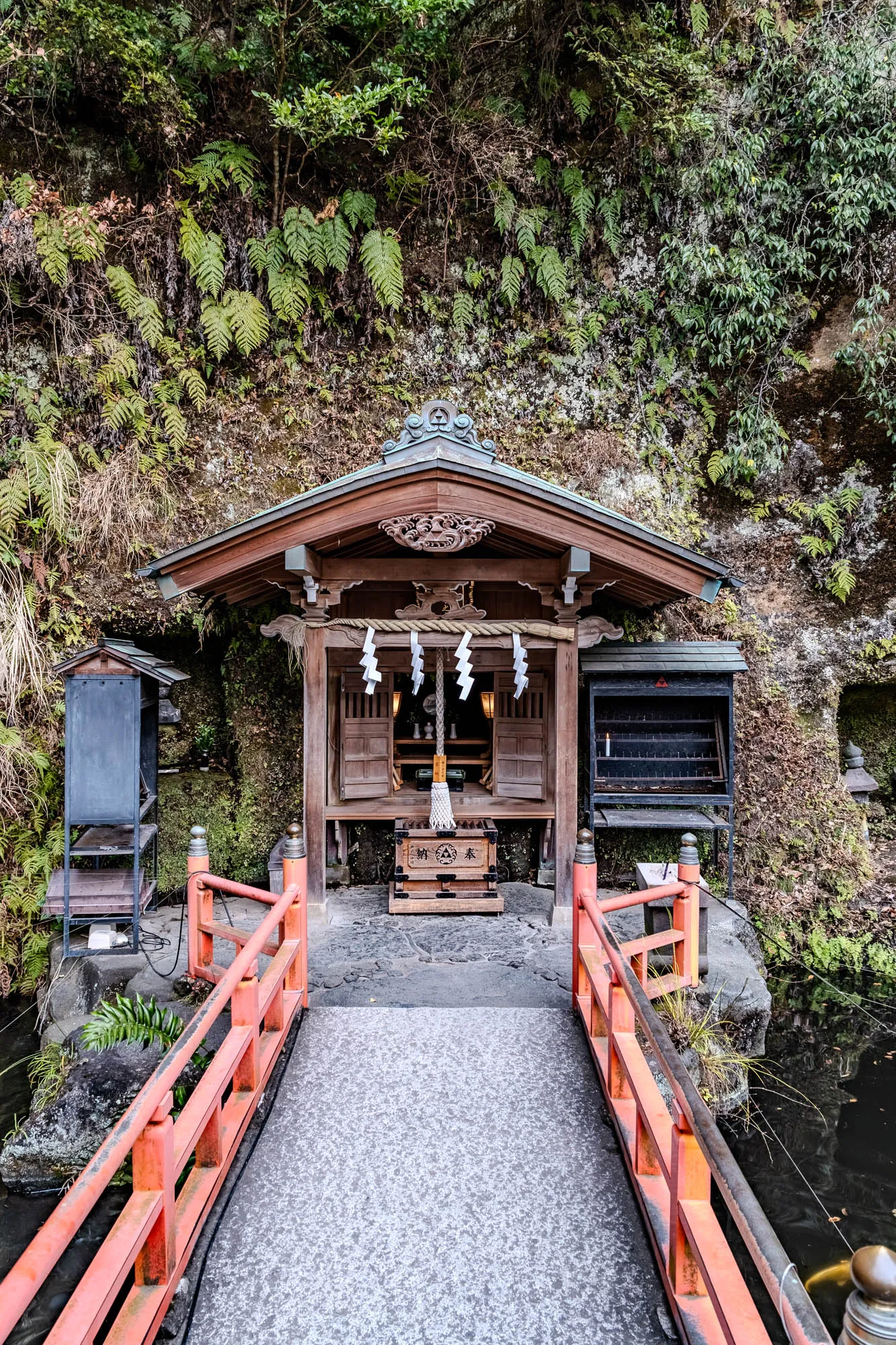 A small, wooden shrine with a red roof sits before a rock face covered in lush greenery. The shrine is adorned with intricate carvings and white paper streamers. A wooden box sits on the floor in front of the shrine. Two black metal stands with shelves are on either side of the shrine. In front of the shrine, a red wooden bridge with a handrail spans a small pond. The bridge is made of red wood and has several horizontal rails, and it's leading into a small, rocky area. The water in the pond is dark and murky. The surrounding foliage is lush and green, with a variety of ferns and other plants. The whole scene has a sense of tranquility and serenity.