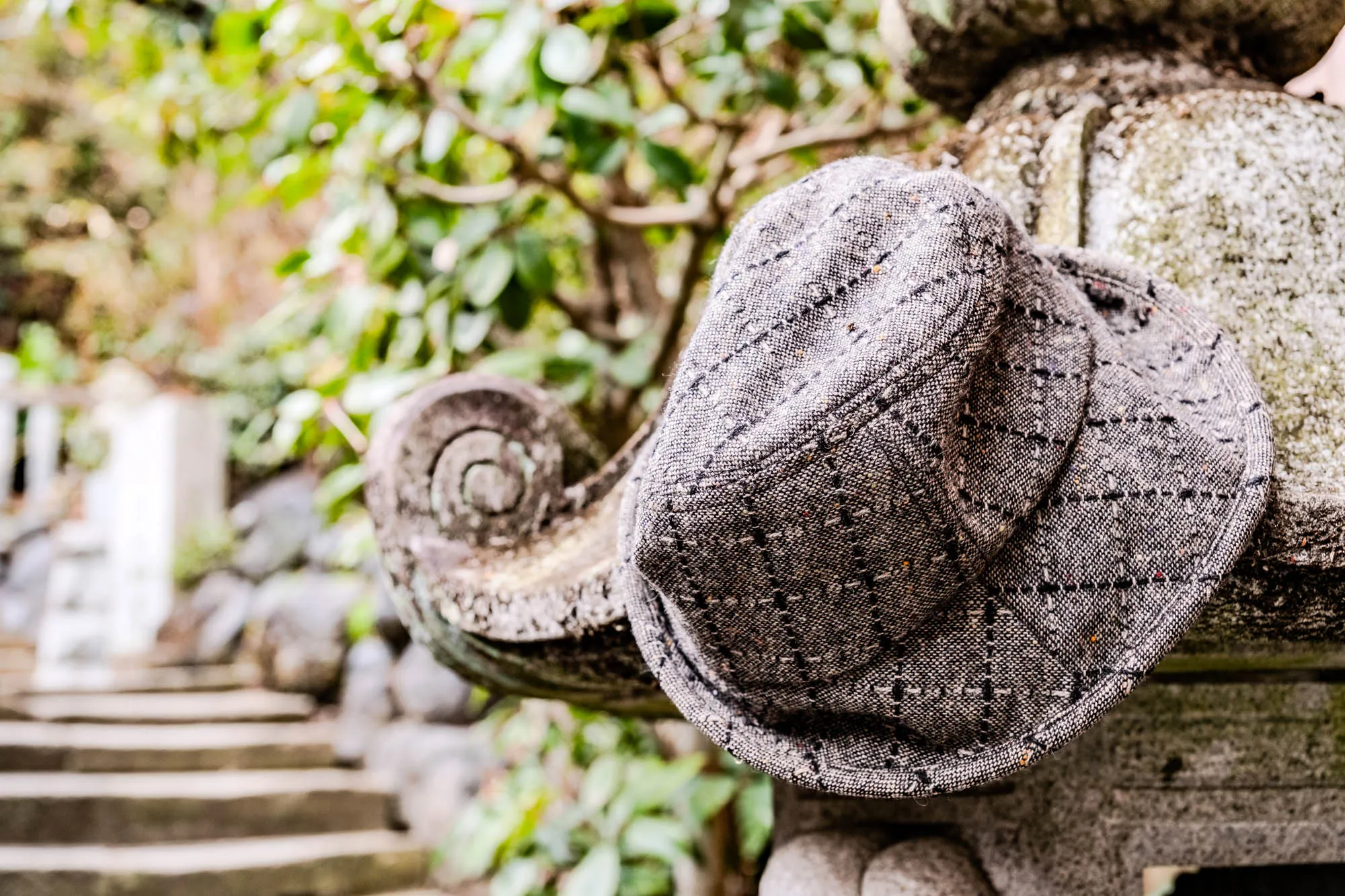 The image shows a close-up of a grey and black hat made of tweed fabric, sitting on top of a stone surface. The hat has a  a woven pattern and looks quite old. The surface of the stone is rough and textured.  The hat is placed on a stone structure, which is likely part of a building or a garden ornament.  In the background, you can hear the sounds of nature, with the rustling of leaves from a bush and perhaps the chirping of birds. There is also a set of stairs in the distance, made of stone, and a hint of white wall in the very distance.  The overall atmosphere of the image is one of tranquility and peace. 
