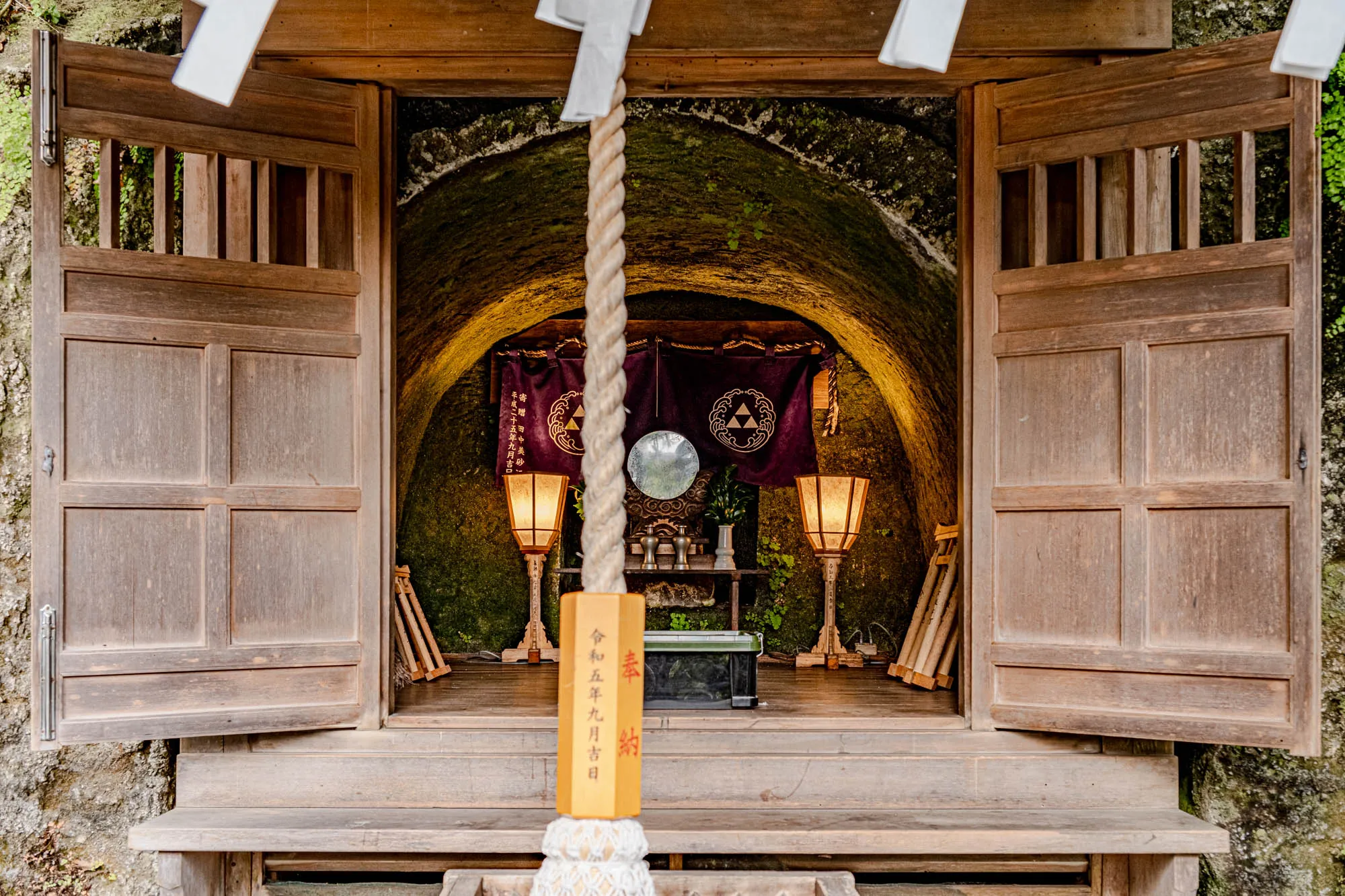 The image shows a small shrine with two wooden doors open revealing the interior. The doors are made of wood and have a grid pattern at the top.  The shrine is located within a stone cave. There is a wooden platform inside with a rope hanging down from the ceiling. The rope has a wooden tag attached to it. The tag has Japanese characters that translate to "Heisei 25 September" and "Reiwa 5 September." The interior of the cave is lit by two lamps with tan shades and there is a circular mirror hanging on the back wall above a small table. Two dark purple cloth banners are hanging next to the mirror and have decorative symbols with writing on them. There are a few small vases on the table along with some other items.  The stone cave is covered with moss and plants and vines. The ground outside the shrine is made of stone and there is a ledge in front of the shrine. The shrine is located in a Japanese forest.  