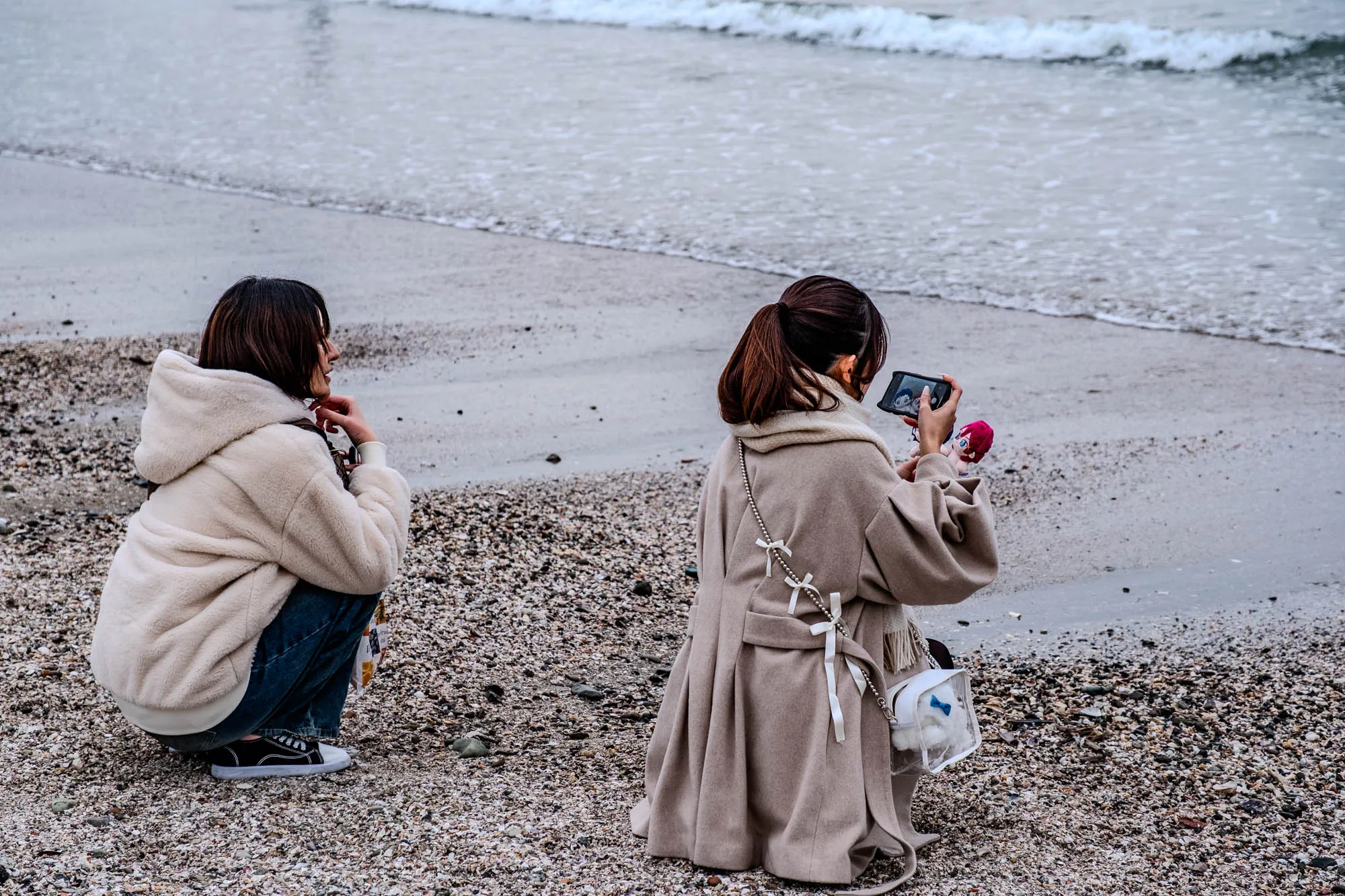 The image shows a beach with two women sitting on the shore. The woman on the left is wearing a white hooded jacket, blue jeans, and black sneakers. She has dark brown hair and is looking to the side. The woman on the right is wearing a beige coat with white bows, a light brown scarf, and a clear purse with a blue bow on it. She has dark brown hair pulled back into a ponytail and is holding a black phone in her hand.  The phone is taking a picture of a small stuffed animal that is pink and blue with a blue bow. The background is a sandy beach with the ocean behind it. The waves are lapping on the shore.