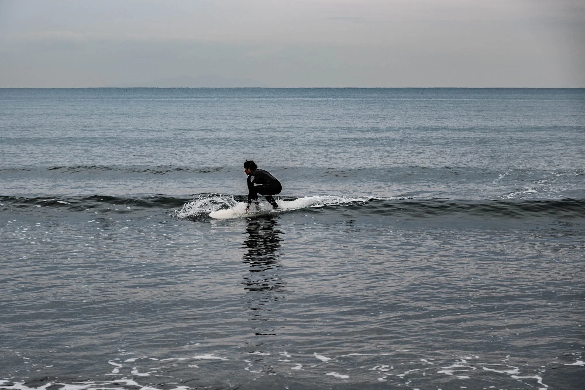 The image shows a surfer riding a wave. They are wearing a black wetsuit and are paddling with their hands in the water. The wave is breaking behind them, creating a white crest. The ocean is a dark blue color, and the sky is a light gray. The surfer's silhouette is clear against the water. There is a faint reflection of the surfer in the water.