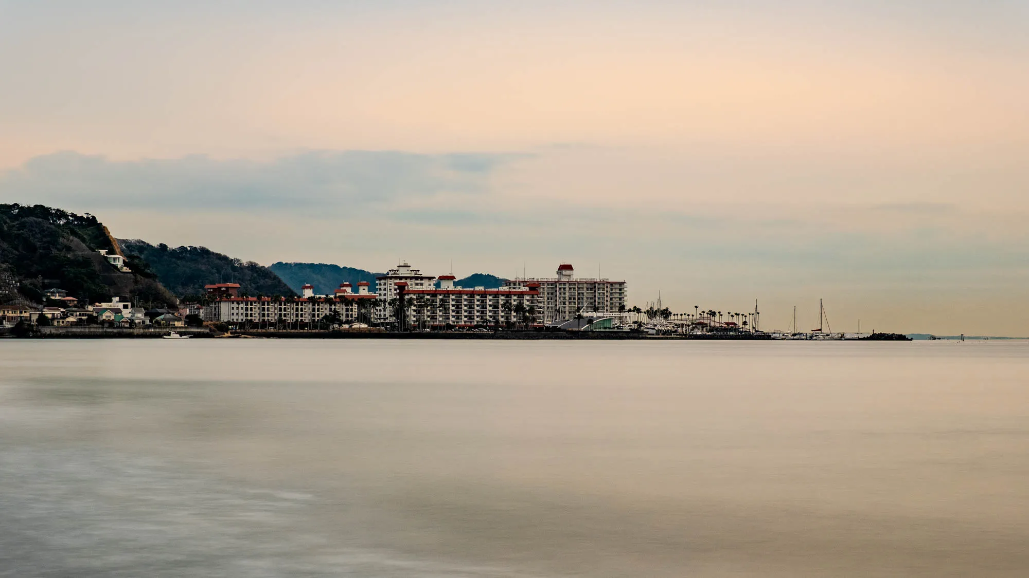 The image shows a distant view of a large hotel complex built on the waterfront. The hotel is white with red accents, with several stories. There are palm trees in front of it, and a marina with boats beyond. There are several small houses in the foreground, set on a small hill.  There is a soft, blurred shoreline and calm water in the foreground. The sky is a light blue and pink, with light clouds. The overall scene gives a sense of peaceful quietude.  
